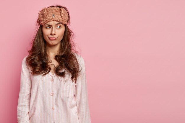 Photo d'une jeune femme détendue et réfléchie a les cheveux bouclés, porte un masque pour les yeux, une combinaison de nuit, concentrée sur le côté, pose sur un mur rose avec un espace libre pour votre contenu promotionnel. Concept de l'heure du coucher