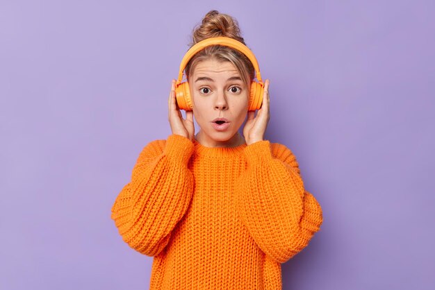 La photo d'une jeune femme européenne choquée retient son souffle ne peut pas croire que ses propres yeux portent un pull ample tricoté écoute de la musique via des écouteurs sans fil pose à l'intérieur sur fond violet Concept Omg