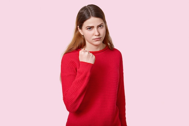 Photo d'une jeune femme européenne en colère triste aux cheveux noirs et raides, montre le poing devant la caméra, vêtue d'un pull rouge, pose sur un mur rose, exprime des menaces, des reproches et promet de se venger.