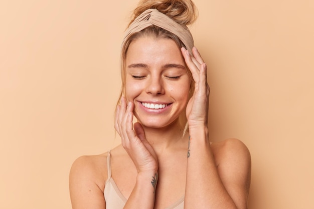 Photo d'une jeune femme européenne heureuse garde les yeux fermés touche le visage doucement a une beauté naturelle une peau propre et saine porte un bandeau a des cheveux peignés isolés sur fond beige. Notion de bien-être