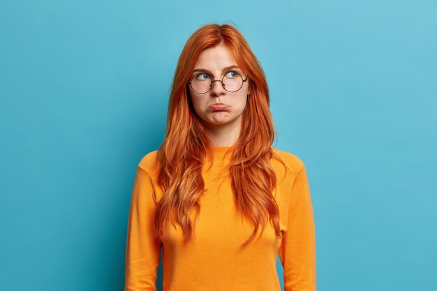 Photo d'une jeune femme européenne rousse offensée a un regard maussade déçu porte un sac à lèvres et détourne son mécontentement en entendant des mots désagréables porte des lunettes rondes, un pull décontracté.