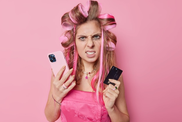 Photo d'une jeune femme irritée fronce les sourcils, serre les dents, applique des bigoudis, tient un téléphone portable et paie en ligne par carte de crédit, porte une robe a une expression agacée isolée sur fond rose