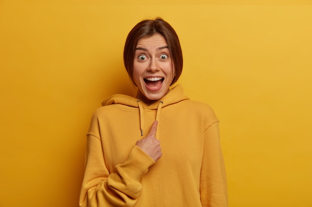 La photo d'une jeune femme joyeuse et excitée surprise se montre elle-même, ne croit pas en être choisie comme leader, porte un sweat-shirt décontracté, isolée sur un mur jaune. Mise au point sélective. Qui, moi?