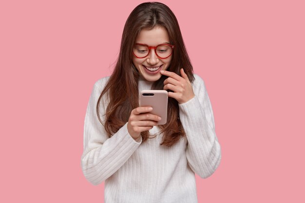 Photo d'une jeune femme joyeuse avec une expression heureuse, tient un téléphone portable, vérifie les actualités des réseaux sociaux en ligne, utilise l'application, porte des lunettes et un pull blanc