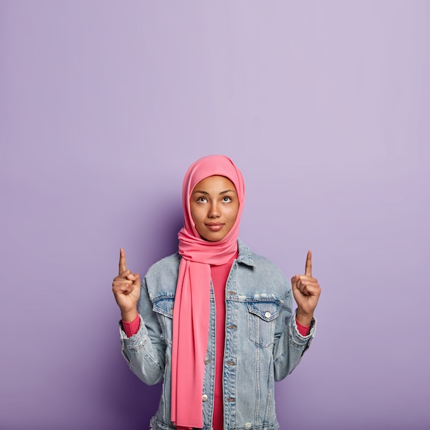 Photo d'une jeune femme musulmane sérieuse pointe vers le haut avec les deux doigts avant, porte un foulard rose, porte une veste rose et en jean, isolée sur un mur violet. Personnes, publicité et promotion.