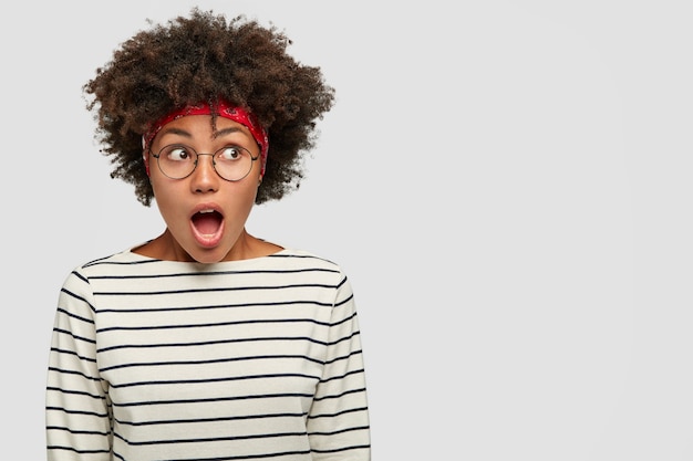 Photo gratuite photo d'une jeune femme noire stupéfaite étonnée ouvre largement la bouche, regarde avec stupéfaction de côté