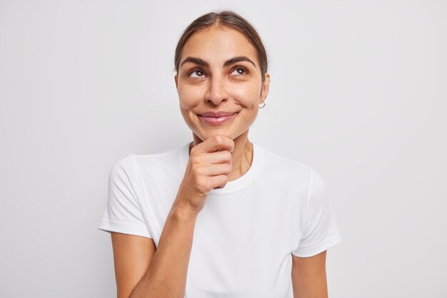 La photo d'une jeune femme ravie et rêveuse tient le menton concentré au-dessus et pense à de nouvelles idées commerciales pour le démarrage porte un t-shirt décontracté isolé sur un mur de studio blanc