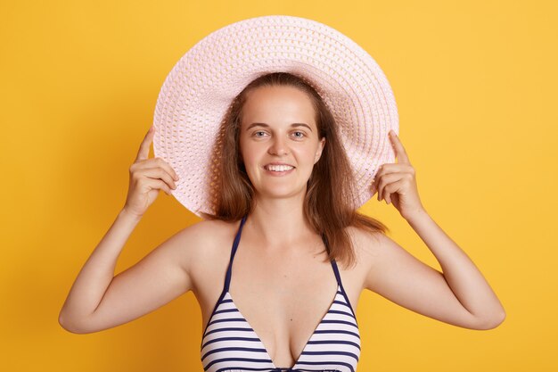 Photo de jeune femme à la recherche agréable en bikini rayé et chapeau de paille, regarde avec une expression heureuse