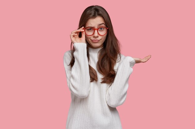 Photo d'une jeune femme sérieuse mécontente regarde scrupuleusement à travers ses lunettes, garde la paume levée, porte un pull surdimensionné