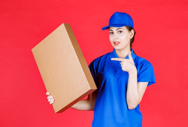 Photo d'une jeune femme en tenue bleue pointant une boîte en carton sur un mur rouge.