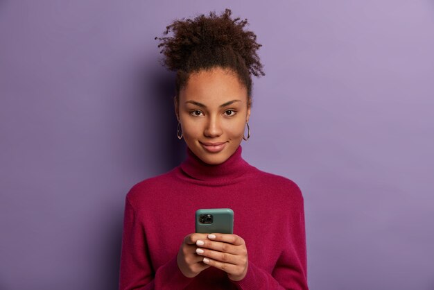 Photo d'une jeune fille du millénaire à la peau sombre et heureuse tient un téléphone mobile, attend un appel, envoie des SMS avec un ami dans un chat en ligne, utilise une application spéciale, appuie sur l'écran du smartphone, vérifie le fil d'actualité, navigue sur Internet