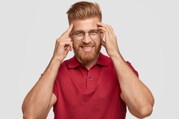 La photo d'un jeune hipster aux cheveux roux joyeux a une mauvaise mémoire, garde les mains sur les tempes, essaie de se souvenir de quelque chose, a un sourire amical, porte une tenue décontractée, se tient seule contre un mur blanc.