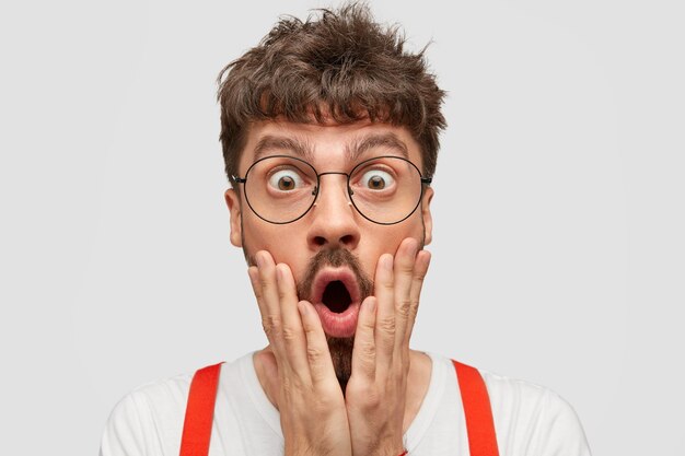 Photo de jeune hipster caucasien étonné garde les yeux ouverts, tient les mains sur les joues
