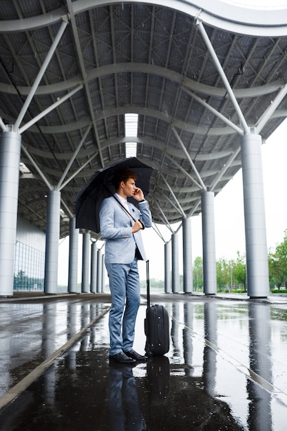 Photo de jeune homme d'affaires aux cheveux roux tenant un parapluie noir sous la pluie et parler au téléphone