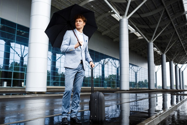 Photo de jeune homme d'affaires aux cheveux roux tenant un parapluie noir et une valise sous la pluie à l'aéroport