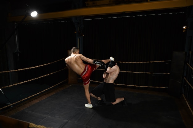 Photo de jeune homme athlétique musclé debout torse nu à l'intérieur du ring de boxe et coups de pied adversaire masculin méconnaissable dans son visage. Concept de personnes, sports, détermination, compétition et rivalité