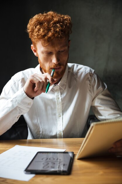 Photo d'un jeune homme barbu à tête de lecture en chemise blanche, lisant des notes, assis à une table en bois