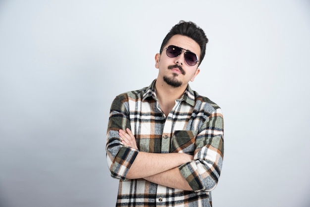 Photo de jeune homme à lunettes noires debout sur un mur blanc.