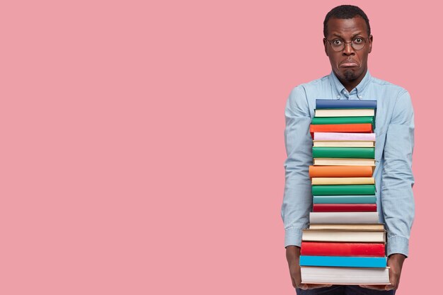 Photo d'un jeune homme noir mécontent tient une pile de manuels, mécontent d'étudier, porte un sac à lèvres, porte des vêtements formels