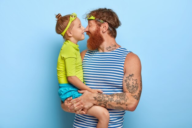 Photo d'un jeune homme rousse barbu porte une petite fille, se touche le nez et a des expressions joyeuses, porte des lunettes, vient à la piscine pour passer la journée activement, exprime son amour l'un à l'autre.