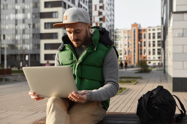 Photo d'un jeune homme sérieux et concentré avec du chaume surfer sur Internet, à l'aide d'une connexion sans fil 4g sur un ordinateur portable. Freelance barbu travaillant à distance sur un gadget électronique générique à l'extérieur dans un paysage urbain