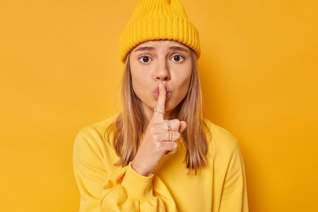 La photo d'une jeune mannequin inquiète fait un geste de chut appuie l'index sur les lèvres dit de se taire demande de garder le silence vêtu d'un pull décontracté et d'un chapeau isolé sur un jaune vif