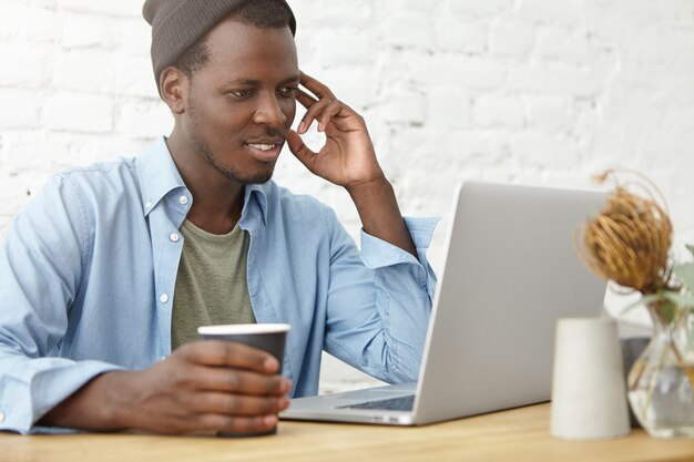 Photo de jeune programmeur afro-américain à la mode, blogueur, pigiste ou startupper à l'aide d'un ordinateur portable à l'espace de coworking pour le travail à distance et boire du café, ayant un regard heureux