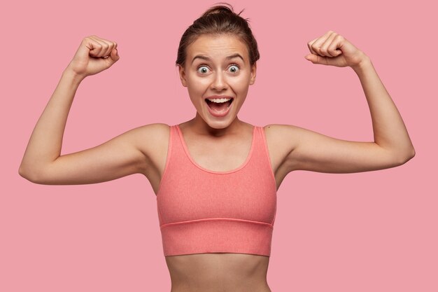 La photo d'une jeune sportive forte et joyeuse montre que les biceps travaillent sur les muscles dans la salle de sport, porte un haut décontracté d'un ton avec un mur, a un corps sain et parfait, étonné. Concept sportif