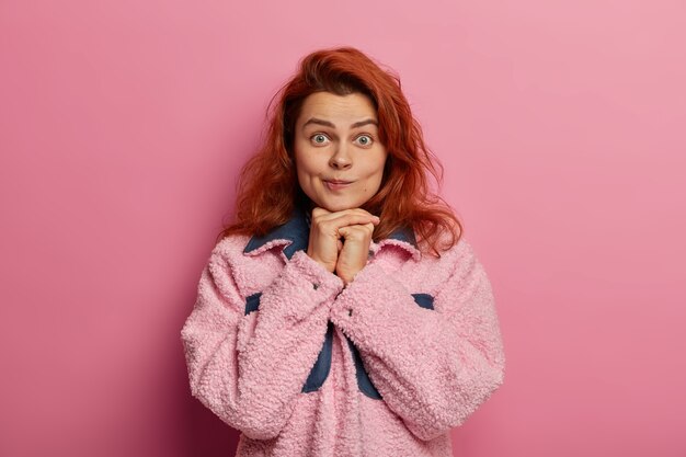 Photo d'une jolie femme aux cheveux roux naturel, garde les mains sous le menton, porte un sac à lèvres, porte un manteau rose, regarde avec émerveillement, dit que je ne crois pas