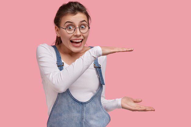 Photo de jolie femme européenne fait un geste de taille, montre la hauteur de l'objet, sourit largement, a une énorme quantité de cadeaux