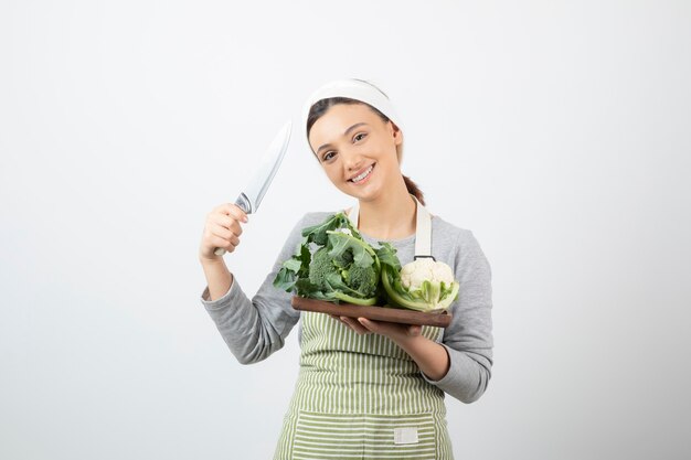 Photo d'une jolie femme souriante tenant un couteau avec une assiette en bois de choux-fleurs