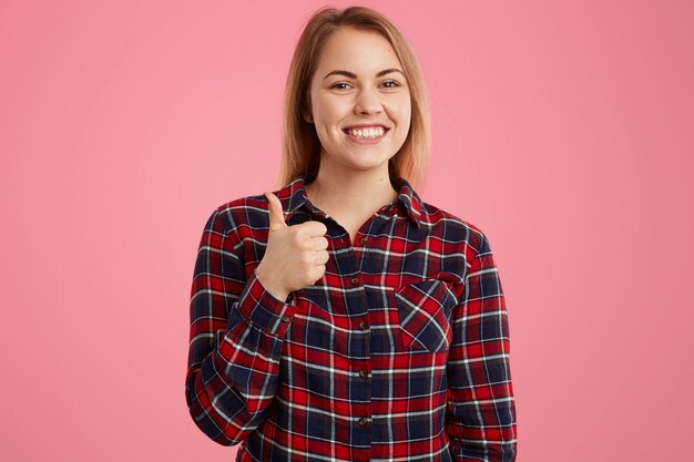 Photo de jolie femme avec un sourire à pleines dents, vêtue d'une chemise à carreaux, garde le pouce levé, donne son approbation à quelque chose