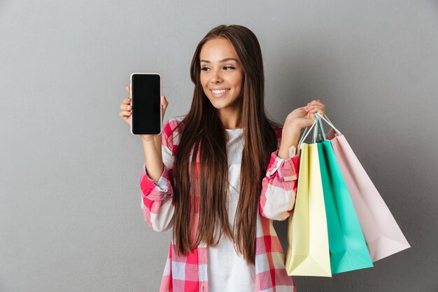 Photo de jolie femme tenant des sacs à provisions, montrant un écran mobile vierge