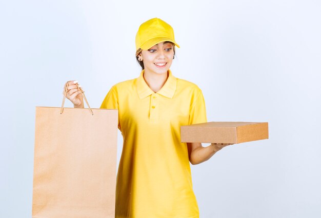 Photo d'une jolie femme en uniforme jaune tenant une boîte et un sac en papier kraft vierge marron.