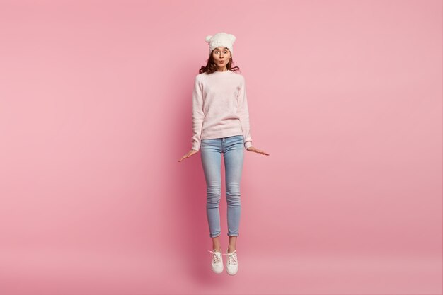 Photo d'une jolie fille saute dans les airs contre l'espace rose, porte un chapeau, un pull, un jean et des baskets, garde les lèvres pliées