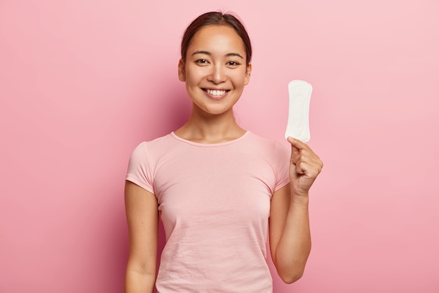 Photo de jolie jeune femme d'aspect asiatique, tient une serviette hygiénique propre, satisfaite de sa qualité, utilise un produit intime pendant les règles ou les règles, isolé sur un mur rose