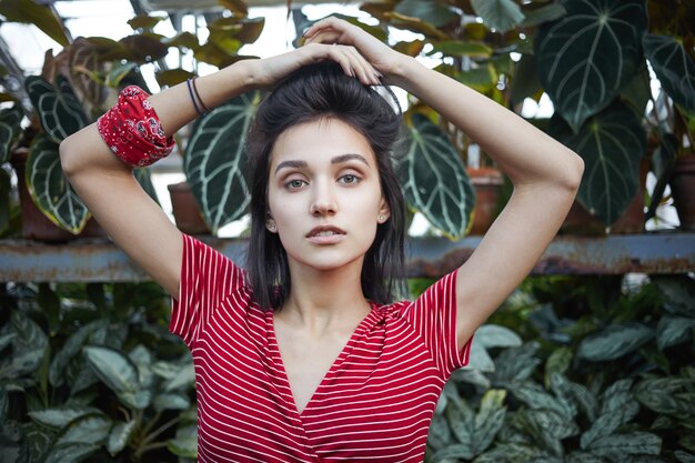 Photo de jolie jeune femme brune en robe élégante posant sur fond de plantes vertes, croisant les mains au-dessus de sa tête et regardant la caméra avec une expression faciale sérieuse et confiante