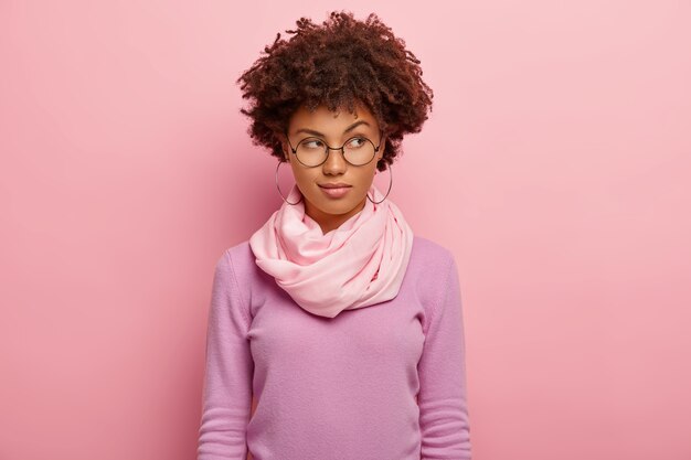 Photo de jolie jeune femme a une expression pensive, soulève les sourcils, regarde de côté, porte des lunettes, un pull violet et un foulard en soie