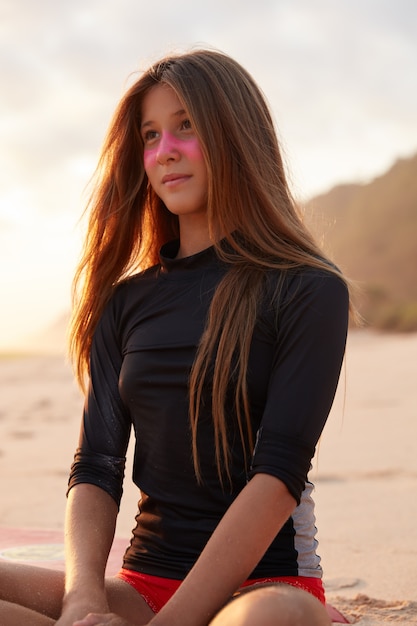 Photo d'une jolie jeune femme rêveuse habillée en maillot de bain, a un masque de zinc rose, s'assoit les jambes croisées sur la plage, pense à quelque chose, se concentre à distance. Concept de personnes, de style de vie et de sport extrême