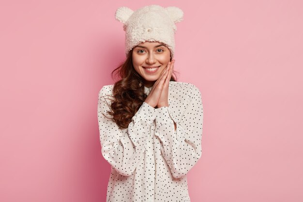 Photo de jolie jeune femme avec un sourire sincère à pleines dents, garde les mains près du visage, se sent heureuse et ravie, porte un chapeau d'hiver chaud drôle avec des oreilles, démontre une nouvelle marque de vêtements cette saison