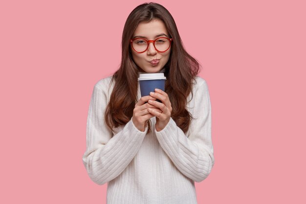 Photo d'une jolie petite amie a l'intention de faire quelque chose, regarde scrupuleusement, porte des lunettes pour une bonne vision, boit du café dans une tasse en papier