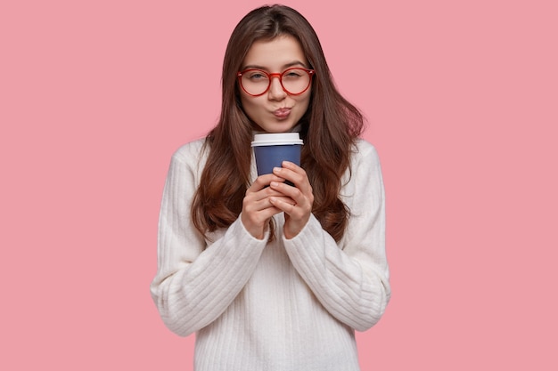 Photo d'une jolie petite amie a l'intention de faire quelque chose, regarde scrupuleusement, porte des lunettes pour une bonne vision, boit du café dans une tasse en papier