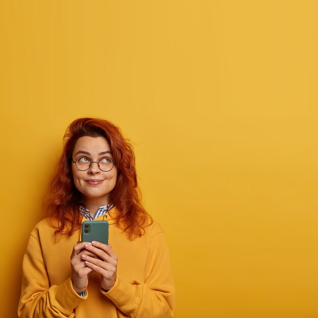 Photo de joyeuse fille millénaire aux cheveux roux utilise un téléphone portable pour envoyer des messages texte, pense au contenu du message pour l'amant