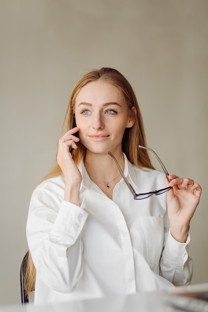 Photo d'une joyeuse jeune femme d'affaires blonde au bureau à l'intérieur de travailler avec un ordinateur portable et un téléphone mobile.