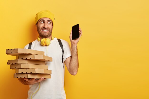 Photo d'un livreur perplexe porte des boîtes à pizza, tient un smartphone moderne, reçoit de nombreux appels et commandes en une seule fois, porte une tenue décontractée, se tient contre le mur jaune