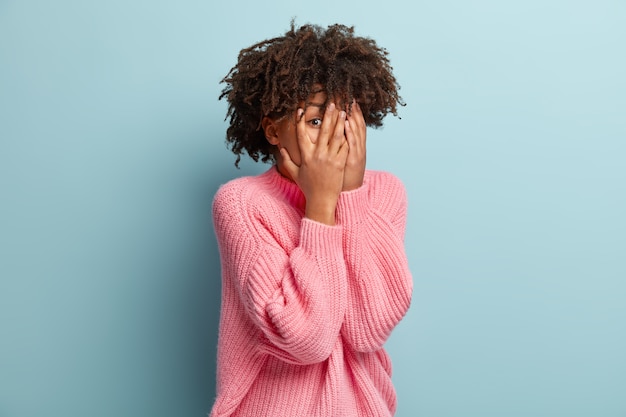 Photo gratuite photo d'un mannequin féminin craintif et embarrassé qui couvre le visage à deux mains, regarde à travers les doigts, observe quelque chose d'effrayant