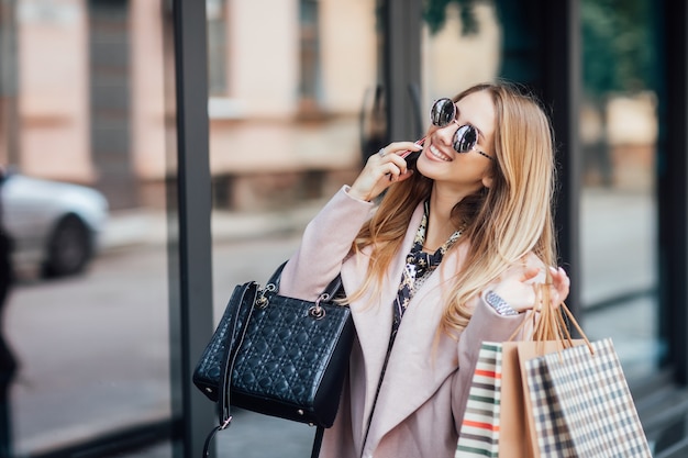 Photo de mode d'une jeune femme blonde élégante marchant dans la rue, portant une tenue tendance, tenant des sacs à provisions et parlant par téléphone.