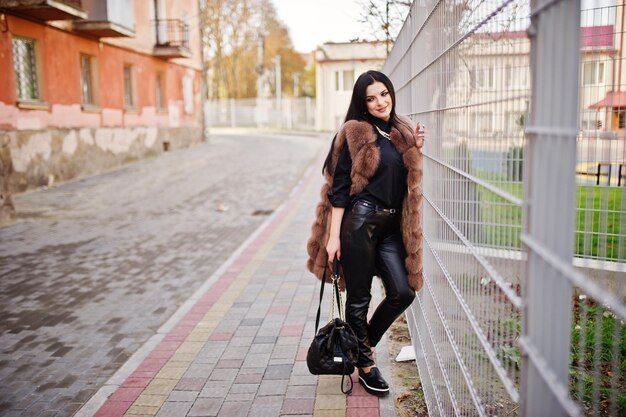 Photo de mode en plein air d'une magnifique femme sensuelle aux cheveux noirs dans des vêtements élégants et un luxueux manteau de fourrure sans manches contre une clôture de fer à la ville d'automne