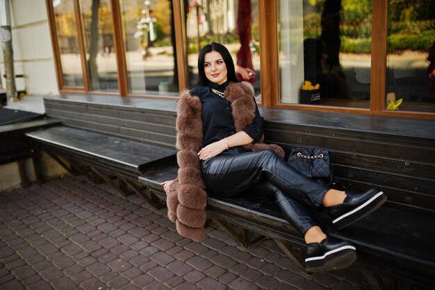 Photo de mode en plein air d'une magnifique femme sensuelle aux cheveux noirs dans des vêtements élégants et un luxueux manteau de fourrure sans manches et avec un sac à dos assis sur un banc à la ville d'automne