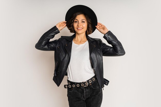 Photo de mode en studio d'une femme brune élégante en chapeau noir et veste en cuir posant sur blanc